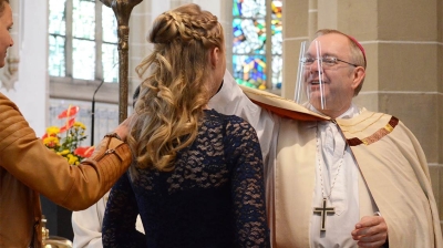 Der Pastor bei der Firmung eines Mädchens. Hinter dem Mädchen steht die Firmpatin und hält die Hand auf die Schulter des Firmlings