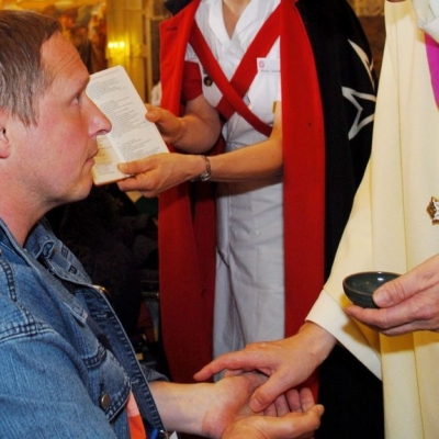 Kranker Mann im Rollstuhl schaut zum Pastor hinauf welcher ihm die Krankalbung gibt. Daneben hält eine Frau ein Gebetbuch offen in der Hand